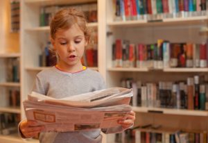 Little girl stands against the backdrop of bookshelves read newspaper